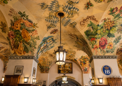 Techo pintado de la Hofbräuhaus de Múnich, con motivos florales, escudos bávaros, lámparas colgantes y decoración histórica.