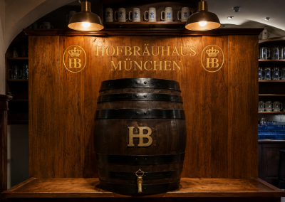 Barril de cerveza de la Hofbräuhaus de Múnich, situado frente a un panel de madera con jarras y el emblema de la cervecería.