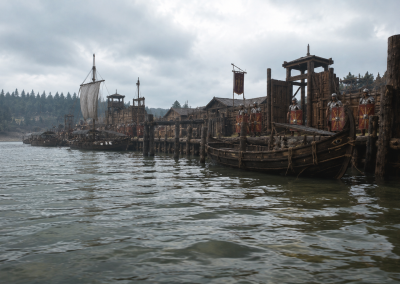 Puerto fluvial romano de Colonia Agrippina, con barcos amarrados, muelles de madera, soldados romanos y un paisaje germánico al fondo.