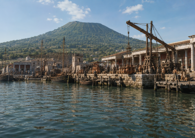 Puerto romano de Pompeya con barcos amarrados, grúas de madera, ánforas, edificios portuarios y el Vesubio al fondo.