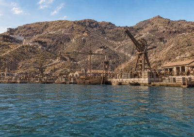 Puerto romano de Portus Magnus con barcos, grúas de madera, muelles de piedra y montañas áridas al fondo.