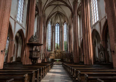 Interior de la Nikolaikirche con nave gótica de ladrillo, columnas altas, bóvedas, bancos de madera, vidrieras y altar al fondo.