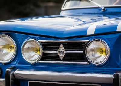 Detalle frontal de un Renault 8 TS azul, con faros redondos, parrilla cromada, franjas blancas y logo histórico de Renault en el centro.